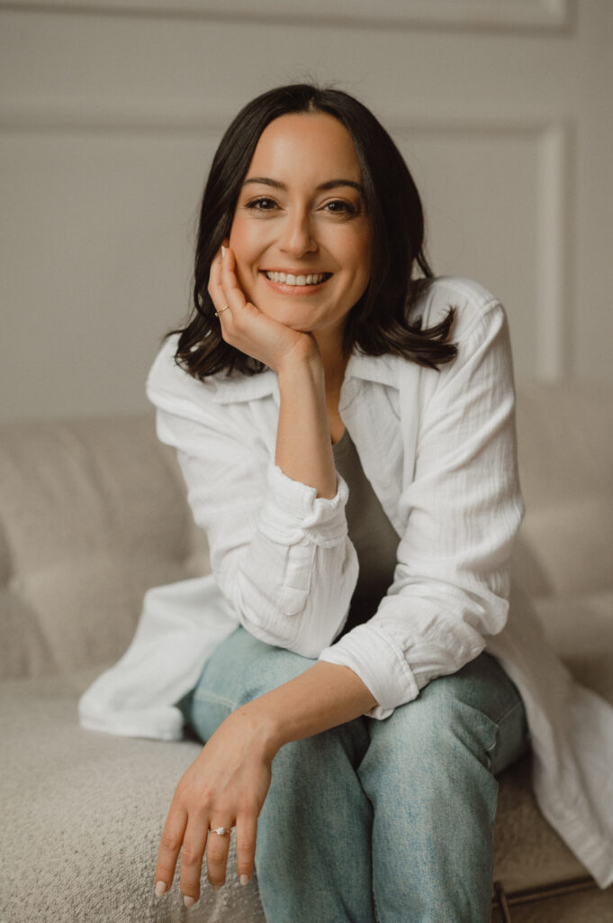 Vickie smiling and sitting casually on a neutral-toned couch, representing a friendly and approachable web designer for wellness professionals.