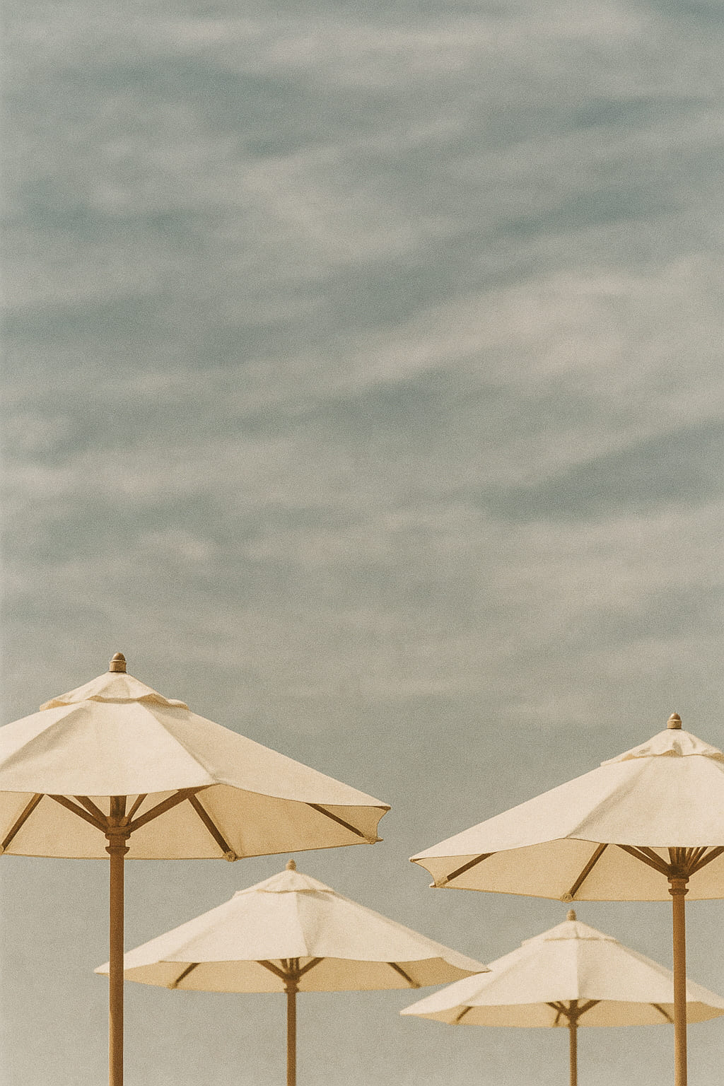 Cream-colored beach umbrellas under a soft, pale blue sky with wispy clouds — relaxed and open summer vibes.