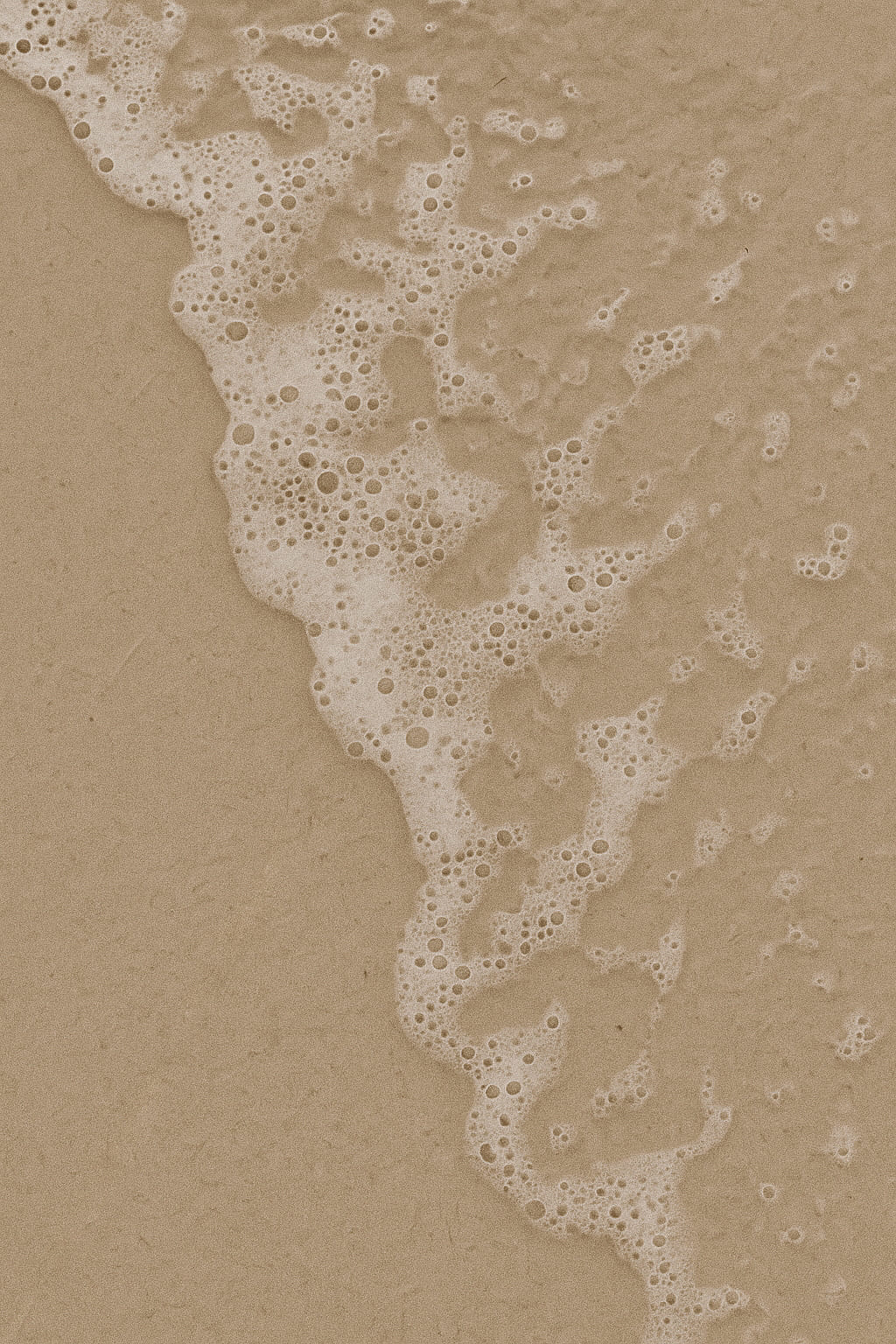 Close-up of foamy ocean water gently washing over smooth beige sand, capturing a calm and minimal summer shoreline moment.
