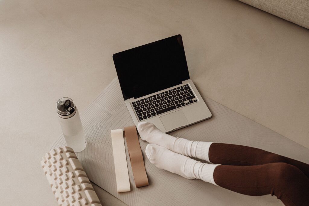 Person sitting on a yoga mat with a laptop, resistance bands, foam roller, and water bottle—representing a calm, at-home wellness setup for virtual sessions.
